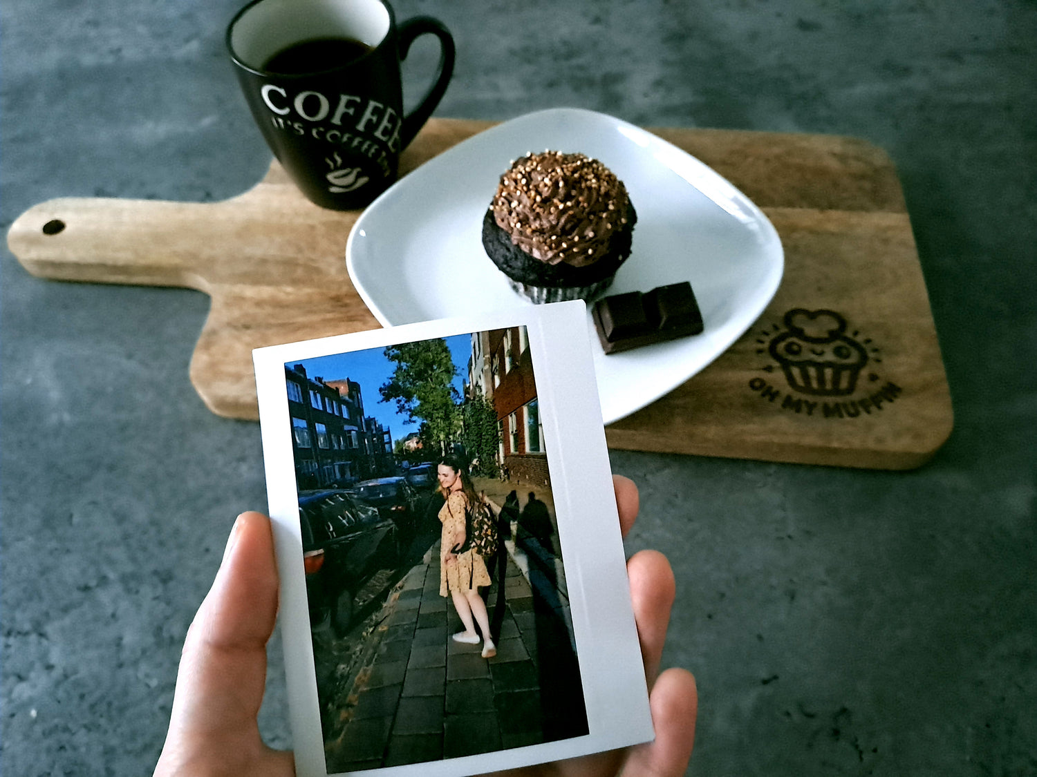 A cup of coffee near a vegan chocolate muffin put on a wooden board with a picture in from of them